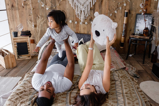 Positive young mixed race family cute young mother African American dad and charming smiling daughter have fun on the bed on Friday evening. Family time concept