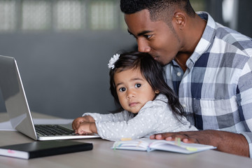 Young caring positive african-american father holding charming mixed race daughter in his arms pushing buttons of laptop while sitting at table with notebooks. Concept of combining business and family