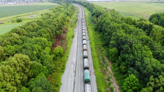 Aerial shot of freight train that travels by rail. Train carries goods and raw materials for heavy industry. Railways are surrounded by trees and endless green fields. Taken by drone at sunset