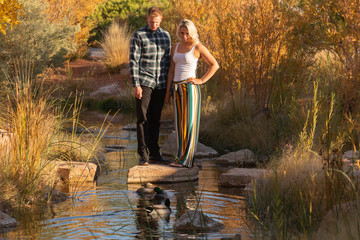 Horizontal image of a young couple standing on a stepping stone in a small stream with autumn foliage behind them watching some ducks swim in the water.