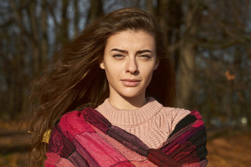 A feminine lady frowns from the sun. Portrait of young woman with brown hair, green eyes wearing pink sweater walking in colorful forest or Park. Girl looking into camera has natural beauty.