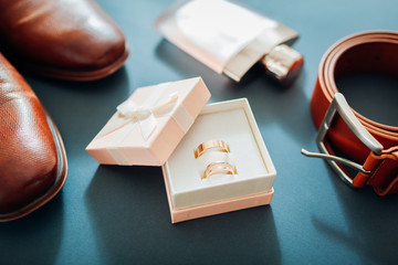 Groom's wedding day accessories. Brown leather shoes, belt, perfume, golden rings on grey background. Male fashion