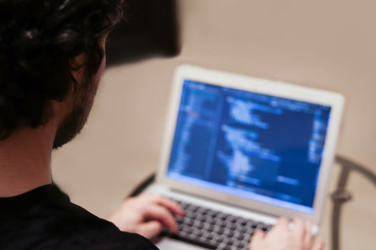 Young man is developing complex software on laptop on blurred background. View from the back without a face. Freelance and programming concept. Program code on screen