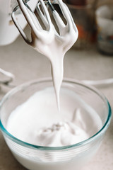 Baking concept. Whipped cream of egg whites for perfect peaks in a glass bowl, with a mixer on a marble white table. Meringue cookie recipe.