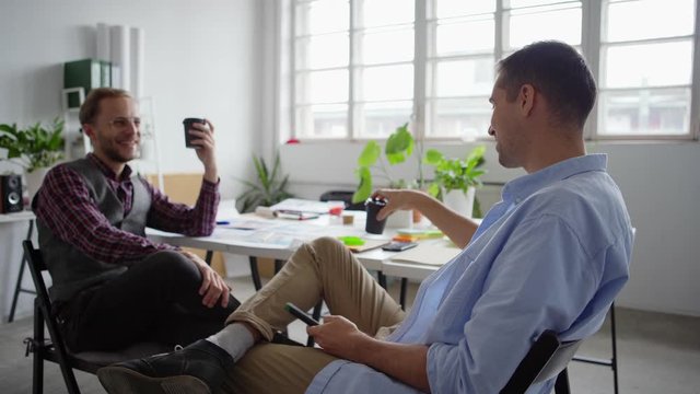 Side view of two cheerful young entrepreneurs sitting on chairs with legs crossed at knee and enjoying talking over coffee in office