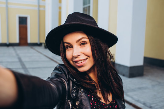 Close-up Fashion Woman portrait of a young girl posing in a city in Europe. Smiling pretty brunette woman in fashionable black hat and leather jacket outdoors. spring walk around the city