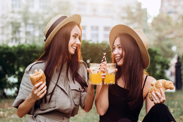 Two cute brunette girlfriends in straw hats are sitting in the park, have a summer picnic in the park. girls drink orange juice and eat croissant outdoors. Close Up portrait of girlfriends at sunset.