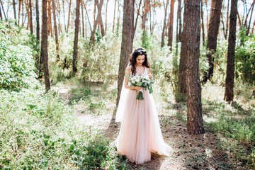 Obraz na płótnie Canvas Beautiful bride in a tender pink dress outdoors. Porter in full growth of a beautiful brunette woman with long dark hair. Girl holds a bouquet. A girl in a long dress walks in a pine forest.