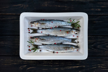 Raw mullet fish with spices and herbs ingredients in white ceramic backing dish on black wooden table ready for cooking. Cooking concept.