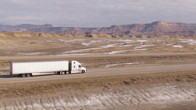 AERIAL: Semi-trailer truck speeds along the motorway running across the desert in Utah. Wintry desert landscape surrounds truck driving down the interstate highway running across rugged US wilderness.