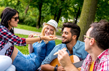 Group of friends in park enjoying spending time together while eating pizza and playing guitar