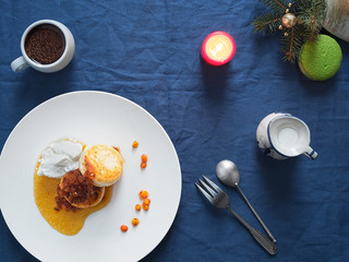 Cheesecakes or curd pancakes with cream and sea buckthorn jam on a white plate, on a blue tablecloth. Vintage gravy boat, espresso. Homemade сottage cheese fritters. Tasty breakfast. Top view.