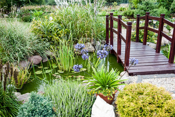 a small stream in a green garden with a wooden bridge and handrails around lush vegetation