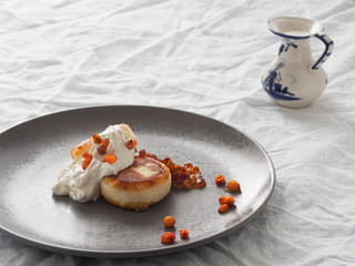 Cheesecakes or curd pancakes with cream and sea buckthorn jam on a gray plate, on a white tablecloth. Vintage gravy boat. Close-up. Homemade сottage cheese fritters. Tasty breakfast. Closeup.
