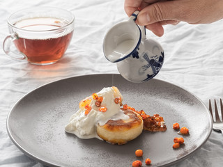 Cheesecakes or curd pancakes with cream and sea buckthorn jam on a gray plate, on a white tablecloth. Vintage gravy boat and cup of tea. Homemade сottage cheese fritters. Tasty breakfast. Closeup.