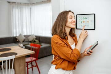 Young woman controlling home with a digital touch screen panel installed on the wall in the living room. Concept of a smart home and mobile application for managing smart devices at home