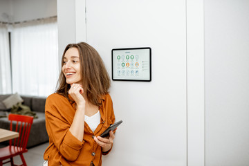 Young woman controlling home with a digital touch screen panel installed on the wall in the living...