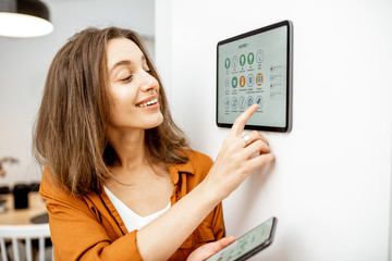 Young woman controlling home with a digital touch screen panel installed on the wall in the living...