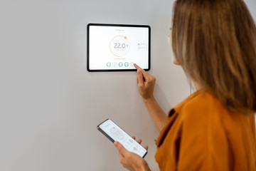 Young woman controlling temperature in the living room with smart phone and digital touch screen panel. Concept of heating control in a smart home - obrazy, fototapety, plakaty