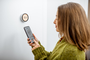 Woman dressed in green sweater regulating heating temperature with a modern wireless thermostat and smart phone at home. Synchronization of thermostat with mobile devices concept - obrazy, fototapety, plakaty