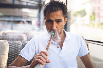 Close-up shot of a stylish adult guy smoking hookah indoor of a cafe