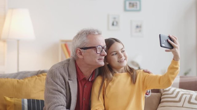 Chest-up shot of smiling 10-year-old Caucasian granddaughter and senior grandfather sitting together on couch at home, girl holding smartphone and taking selfies, then looking at photos on screen