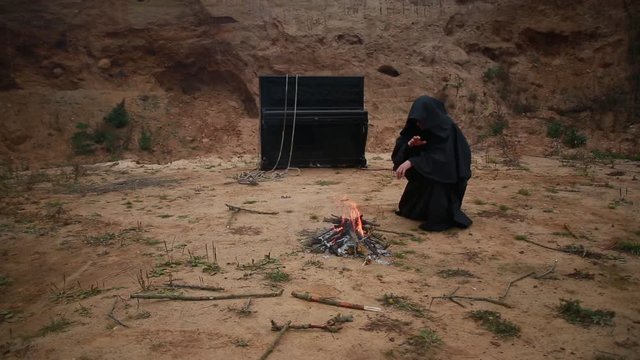 A monk in a black robe with a hood sits on a vacant lot and warms himself by the fire