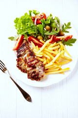 Grilled pork steak with french fries and fresh salad. White wooden background. 