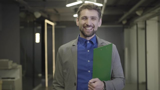Portrait of tired Caucasian male office worker yawning, looking at camera and smiling. Bearded man in formal suit standing with document folder in open space. Management, coworking, overworking.