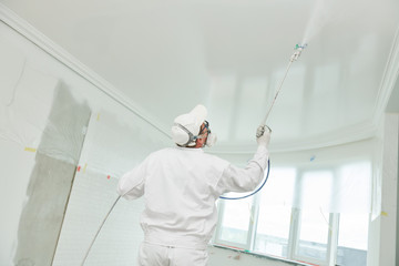 Painter worker with airless painting sprayer covering ceiling surface into white
