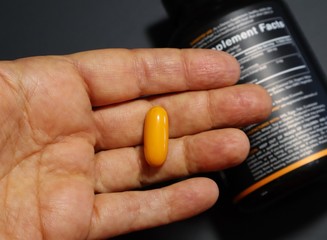 Closeup of a hand holding a turmeric curcumin capsule with blurred supplement bottle label in the background