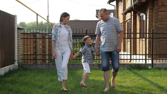 handsome father and pretty young mother hold happy little son hands and walk on green grass against fence slow motion