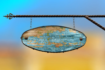 Oval wooden blue sign. An old tattered wooden shopboard with no text hanging on a wrought iron structure. Template on blurred city background.