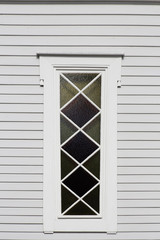 Fototapeta premium Window of traditional wooden rural house. Abstract village architecture fragment featuring narrow stained glass, triangular and rhomboid frames and white wood siding or wall cladding.