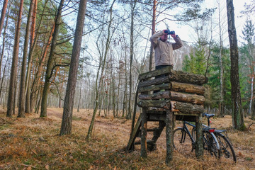 A 59 year old man watches binoculars. Bicycle stands beside a wooden observation tower. Hunting...