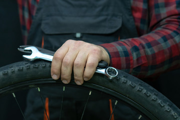bike repair. Bike mechanic in the workshop. A mechanic holds tools and a bicycle wheel in his hand. Hands of the master and keys close-up. A man in a fashionable plaid shirt and work overalls.