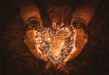 Selective focus on woman hands wearing knitted mittens and holding glowing glass heart shape in winter, with soft fur background with copy space.