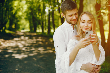 Naklejka premium A beautiful and gentle girl with light hair and a white dress is walking in a sunny summer forest with her handsome guy in a white shirt and dark pants and they use the phone