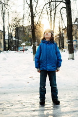 A nine year old school age boy standing in the street in a blue jacket and hat in the backlight looking at the sky. Winter frosty sunset of the day walking 