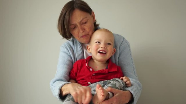 Close portrait of a happy grandmother and her young grandson. The woman holds the child in her arms and tickles his heels, the baby laughs cheerfully. Happy family weekend