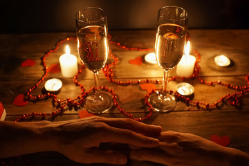 Close-up of a pair of hands on a table with two glasses of champagne, candles, hearts, beads. A romantic couple hold each other's hand during dinner. concept of engagement.