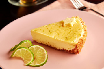 Piece of delicious tart with lime zest and slice of limes on pink plate on dark wooden table, served with cup of herbal tea.