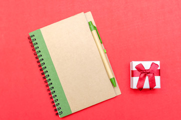 Notepad with pen and gift on red background. Flat position, top view. The concept of the holiday,...