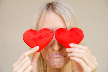 Lifestyle concept: sad, resentful attractive blonde with two small red hearts in her hands, closing her eyes with heart symbols, standing on a light background. Valentine's day