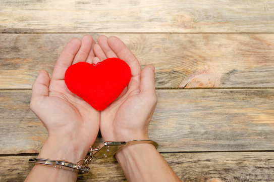 Man's handcuffed hands hold red heart on wooden background, copy space