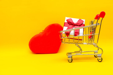 gift box in shopping cart on yellow background next to red hearts, copy space. Concept of gifts, Valentine's day, holiday