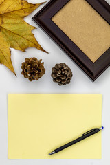 Composition Using Autumn Leaf, Empty Picture Frame, Blank Paper For Text, Black Pencil, and Two Pine Cones on White Background
