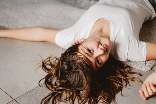 Lifestyle portrait of pretty playful teenager girl with messy hair lying on floor looking at camera with smile. Leisure time at home.  View from above,
