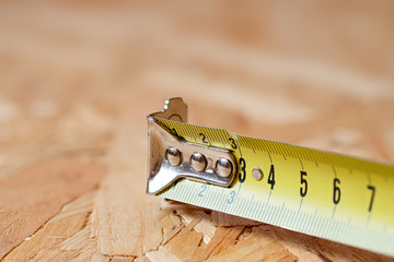 A tip of yellow metal tape measure lying on chip board. Blurred background.