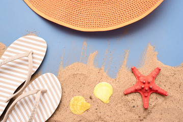 Female hat, seashells, red starfish and flip flops shoes on beach sand over the blue background.